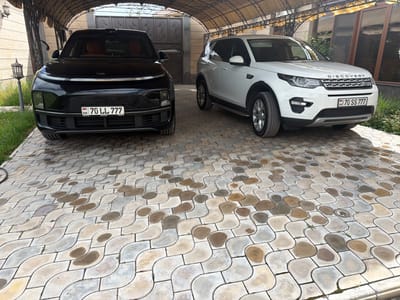 Two cars, a black vehicle with license plate "AM 70 LL 777" and a white "Land Rover Discovery" with license plate "AM 70 SS 777", are parked under a carport on a patterned stone driveway in front of a building. A lamppost stands in the yard on the left. The OCR text identifies "XMOTORS," and the license plates are Armenian. The location is likely in Armenia, potentially in or near the city of Dimitrov, Armenia.