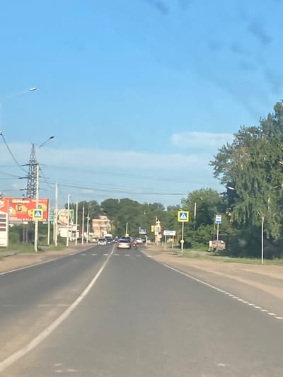 The image shows a road in Rubtsovsk, Russia, with cars driving on it. The road is asphalt and has a white line marking the lane. On the left side of the road, there are trees and buildings. There are also several traffic signs visible, including pedestrian crossing signs and other road signs. One sign in the foreground reads "СВЕТОФОР" which means "traffic light". The sky is blue and it appears to be a sunny day. The image is taken from inside a vehicle, likely while it is moving.