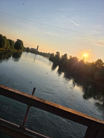The image depicts a tranquil river scene at sunset, likely from a bridge in Boara Pisani, Italy. The sun, a bright orb, is setting on the right side of the frame, casting a warm glow across the sky and reflecting on the rippling water. Trees line the riverbanks, creating a lush, green border. In the distance, a tall, slender building, possibly a church tower or monument, is silhouetted against the sunset. The sky is a gradient of soft blues and oranges, with a faint contrail from an aircraft visible. Two birds are seen flying over the river, adding a sense of movement to the otherwise still landscape. The foreground of the image is dominated by the rustic metal railing of a bridge, providing a clear viewpoint of the scene. The water's surface is disturbed by gentle currents, with reflections of the trees and sky visible.