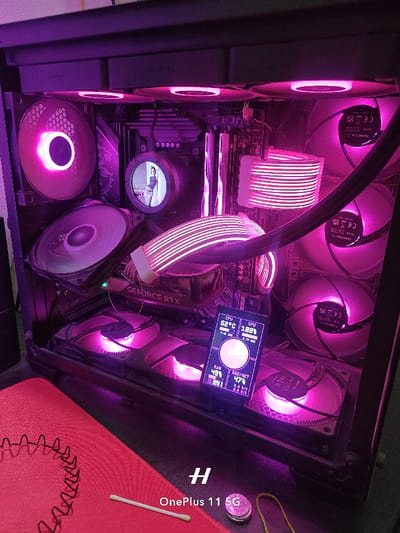 The image shows a close-up view of the inside of a computer case, illuminated with pink LEDs. The case contains numerous fans, a motherboard, and a graphics card. There is a small LCD display attached to the case, showing system information. The environment is a room with a red mat and a piece of string. The image was taken in Rajshahi, Bangladesh, at the coordinates 24.388 latitude, 88.592 longitude. The scene is static, depicting a computer system that is presumably switched on and in operation. There are no people or objects present. No actions or interactions are taking place. The environment is indoors, with a red mat and string visible.