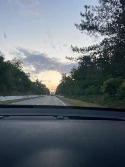 This media file captures a view from inside a vehicle, looking out through the windshield. The scene depicts a road bordered by dense green trees on either side, suggesting a rural or wooded setting. The road itself is paved and appears to be a two-lane highway, with a white line marking the center and the edge. In the distance, a white truck is visible driving on the road.

The sky is a mix of light blue and gray clouds, with a warm glow of sunset or sunrise visible on the horizon. This indicates that the time of day is either dawn or dusk. The overall mood conveyed is one of travel and serene natural beauty.

The foreground of the image is dominated by the interior of the car, specifically the dashboard and parts of the windshield frame. This framing emphasizes the perspective of someone traveling within the vehicle. There are no people or explicit objects identifiable beyond the truck and the natural elements. The location is Hrușova, Moldova, which provides a geographical context to the scene. No text is visible within the media file.