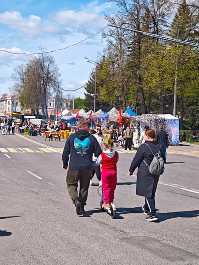 A medium, eye-level shot captures a bustling street scene in Uglich, Russia. A family is walking across the street. The father, wearing a dark hoodie and khaki pants, holds the hand of his child, who is dressed in red and riding a scooter. To their right, a woman in a black coat with a gray backpack walks alongside. In the background, a fair or market is underway, with numerous stalls and people gathered at tables eating. The architecture of Uglich, with its historic buildings, is visible in the distance. The sky is a mix of blue and scattered white clouds. The road is marked with a crosswalk. Trees line the street, adding to the vibrancy of the scene.