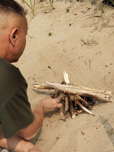 A man is kneeling on a sandy beach, in the city of Bibikha, Russia, tending to a small bonfire made of driftwood. He is wearing a dark green t-shirt and his arms and legs are covered in sand. The bonfire consists of several pieces of dry wood, with flames visible at the base, indicating it is actively burning. In the background, there are patches of dry grass and sparse vegetation, consistent with a beach environment. The sand is light brown and appears somewhat uneven, with small debris scattered across its surface. The lighting suggests it is daytime, with the sun casting shadows on the sand.
