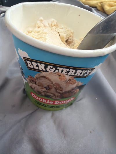 A close-up, eye-level shot shows a pint of Ben & Jerry's Cookie Dough ice cream on a wrinkled, light gray sheet. The ice cream is partially scooped out, with a metal spoon resting inside. The pint's blue exterior features the iconic Ben & Jerry's logo and a cow illustration, as well as the flavor name "Cookie Dough" in red lettering against a green background. The scene is lit with natural light, casting soft shadows. There are no people visible in the frame. The background consists of the sheet, with a hint of a yellow fabric in the upper right corner. The image appears to be taken indoors, possibly in a bedroom or living area in Pentre, United Kingdom, given the domestic setting and the potential for local products.