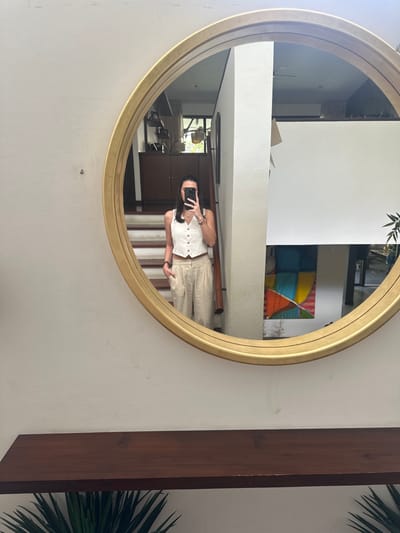 A woman is reflected in a round, gold-framed mirror hanging on a white wall. She is taking a photo of herself with her phone. She wears a white button-down top and beige pants. Behind her in the mirror's reflection are stairs, a colorful painting, and various interior architectural elements. To the right of the mirror is a dark wooden beam with green plants behind it.