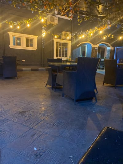 The image depicts an unoccupied outdoor seating area, likely a patio or lounge, located in Igbessa, Nigeria, at night. The ground is paved with dark grey concrete tiles patterned to mimic wood planks and stone pebbles, exhibiting visible wet patches and minimal debris.
The scene features multiple dark-colored, possibly wicker-style, chairs and glass-topped tables. In the mid-ground, a square table with four surrounding chairs is prominent, with another chair to its left. A single chair is situated further left, and additional furniture is partially visible on the right periphery.
In the background, a dark grey building wall dominates the left side, featuring two ornate white-framed windows, each containing multiple panes. Two outdoor air conditioning units are mounted on this wall above the windows. Overhead, string lights with numerous exposed bulbs illuminate the area, intertwined with artificial foliage in various colors, creating a decorative canopy.
To the right, a separate structure with two arched entryways is visible, exhibiting dark, possibly tiled or marble, facings. The word "BAR" is faintly visible above the right archway. The interior beyond these arches appears illuminated with a blue light. No individuals or specific actions are present within the frame.