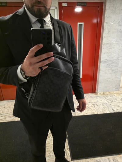 A man dressed in a black suit, white shirt, and black tie is standing indoors, holding a smartphone in his left hand and a black sling bag across his body with his right arm. The sling bag is made of leather and has a patterned exterior. The man has a beard and is looking down at his phone. Behind him, there are red doors with white trim and a sign that reads "2BI49 238A13" in reverse. The floor is a marble pattern. The scene appears to be in an apartment building hallway in Montrouge, France, given the bilingual signage on the door which indicates "ÉTAGES PAIRS" (even floors) in French and the building code.