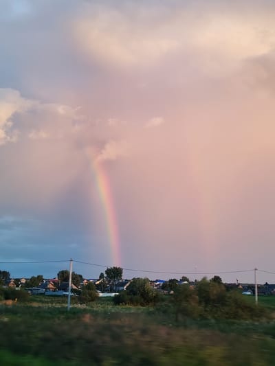 A rainbow arches through a hazy pink sky over a rural landscape, featuring a small village with houses and trees. The lower portion of the image shows a blurry green foreground, suggesting motion, possibly from a vehicle. The scene is set in the village of Bob'ya-Ucha, Russia.