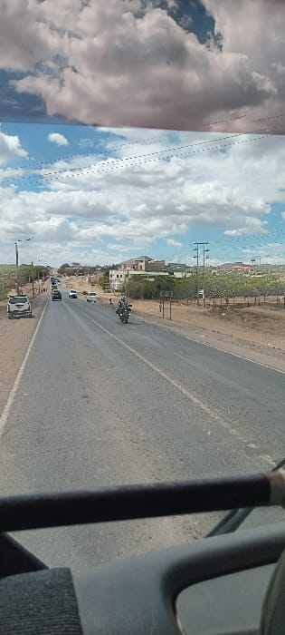 A slightly tilted shot shows an asphalt road winding through a landscape under a partly cloudy sky. Buildings and greenery line the side of the road, accompanied by utility poles. A motorcycle and several vehicles are visible on the road, suggesting traffic. A portion of what appears to be a vehicle interior, specifically a pole and dashboard area, occupies the right edge of the frame, creating a sense of being inside a moving vehicle.