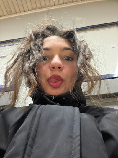 A young woman poses for a selfie, her face tilted towards the camera with her tongue sticking out. She has brown curly hair, a fair complexion, and dark eyebrows. She wears a black jacket, partially visible on the right side of the frame. The background appears to be an indoor space with a tiled wall or surface. The lighting is bright and slightly harsh, possibly fluorescent.