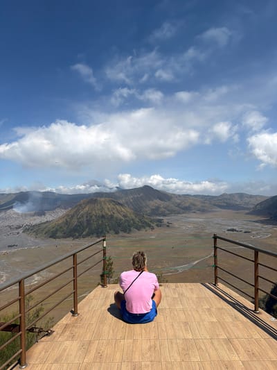 A person wearing a pink shirt and blue shorts is sitting on a wooden platform, facing away from the camera, looking out at a volcanic landscape. The platform has a metal railing. In the background, several volcanoes are visible, with smoke or steam rising from one of them. The sky is blue with scattered clouds. The scene is likely located at a viewpoint in Cemorolawang, Indonesia, overlooking the Bromo Tengger Semeru National Park. The terrain is rugged and mountainous, with a wide, sandy plain in the middle ground.