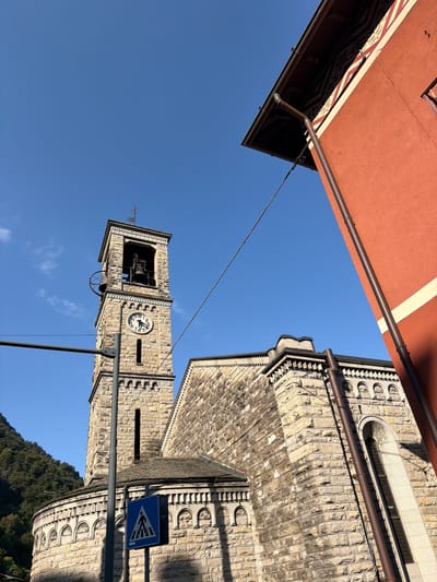The image is an outdoor, low-angle, eye-level shot of a stone church with a prominent bell tower and a portion of a red building to the right. The sky is clear and blue with a few wispy clouds. A pedestrian crossing sign is visible in the foreground. The church features arched windows and decorative stonework. The red building has a dark roof and decorative trim. The scene appears to be in the town of Carate Urio, Italy, given the architecture and setting. There are no people or animals clearly visible in the image.