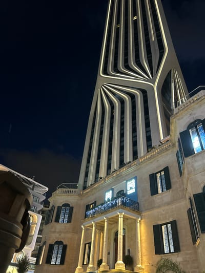 The scene depicts the Mercury Towers complex in Saint Julian's, Malta, observed at night. A tall, slender skyscraper, featuring a light-colored facade with numerous integrated vertical and curved LED light strips, dominates the background. These lights create a distinctive, radial, or fan-like pattern on the lower portion of the tower's structure. In the foreground, a multi-story, traditional-style building constructed from light-colored stone stands brightly illuminated by warm-toned external lights. This building possesses a classical portico entrance with four columns supporting an upper-level balcony. Above the balcony, a luminous sign displays the text "M MERCURY." The balcony railing is adorned with decorative string lights. Multiple windows are visible across the traditional building's facade, some equipped with dark shutters. The sky is dark with minimal cloud cover. A partial view of another modern building is visible on the far left. No individuals or active interactions are present in the image.