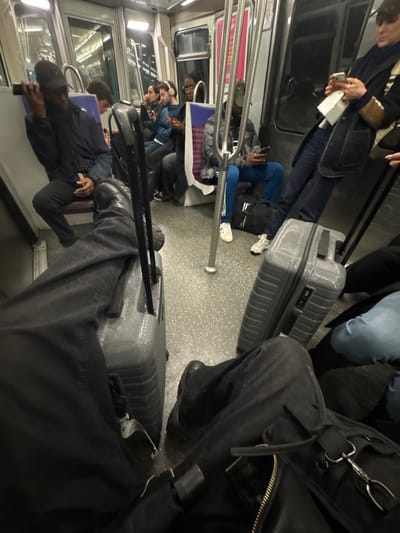 The image depicts the interior of a public transport train carriage, operating within the city of Gennevilliers, France. The perspective is from a seated passenger looking forward into the carriage.
In the immediate foreground, the photographer's legs are visible, clad in dark trousers and dark shoes, with a dark bag resting on their lap. A grey, hard-shell suitcase with an extended handle and textured surface is positioned on the floor between the photographer's feet.
On the left side of the carriage, a male passenger wearing a dark jacket and a black baseball cap is seated, holding a dark mobile phone to his left ear. Behind him, a second passenger with curly hair is visible, looking down at a device. Further along the left side, two more individuals are seated, each viewing a mobile phone; one wears headphones and a blue jacket, the other a dark jacket. A person with a dark head covering is partially visible between these seated individuals.
On the right side of the carriage, a male passenger wearing a grey puffer jacket, a camouflage-patterned baseball cap, and blue track pants with white stripes, is seated directly on a black duffel bag placed on the floor. He is holding and viewing a mobile phone. Another grey, hard-shell suitcase with a combination lock and textured surface is positioned on the floor to his right. Further right, a passenger in a dark coat and blue scarf is standing or leaning, holding a mobile phone and a white document. The lower legs of an additional passenger, wearing light blue jeans, are visible at the far right of the frame.
The train interior features a grey speckled floor, white and light-colored wall panels, silver vertical support poles, and seats with purple and white striped upholstery. Overhead strip lighting illuminates the carriage. Windows show reflections or an indistinct dark exterior. Most visible individuals are engaged with personal mobile devices.