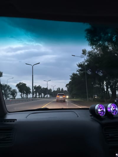 The image depicts a view from inside a car driving on a highway in Достук, Kyrgyzstan. The time of day appears to be dusk, with a cloudy sky and a hint of twilight. The road is paved with a yellow line marking the lanes. On the side of the road, there are several tall streetlights and a line of trees. A white SUV is visible ahead on the highway. The car's dashboard is partially visible on the right side of the frame, featuring two illuminated gauges with purple light. The windshield shows a reflection of trees and the sky, with a large portion of the left side of the frame dominated by a dark, possibly tinted, area of the car interior or a reflection. The overall mood is somewhat subdued, typical of a drive during twilight.