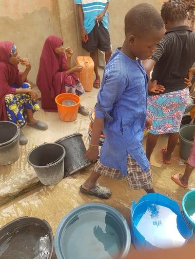 In Dutsinma A, Nigeria, a group of people stand and sit near an assortment of buckets, barrels, and containers, many of which are filled with water. A young boy in a blue shirt and plaid shorts is walking. Several of the individuals are women, some wearing maroon hijabs and colorful patterned dresses. The ground appears to be wet and possibly muddy.