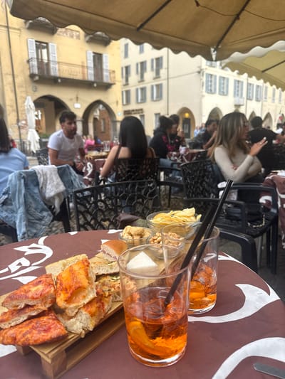 The image depicts an outdoor cafe setting, likely in the city of Pavia, Italy, as indicated by the architecture in the background. Multiple people are seated at tables, engaged in various activities such as using mobile phones and conversing. On one of the tables, two glasses of orange-colored beverages (possibly Aperol spritzes) are visible, along with small bowls containing snacks like chips and peanuts. A wooden tray holding assorted sandwiches and bread is also on the table. The scene is captured under a light-colored awning, with historic buildings featuring arched walkways and multiple windows serving as a backdrop. A casual atmosphere is suggested by the clothing of the individuals present.