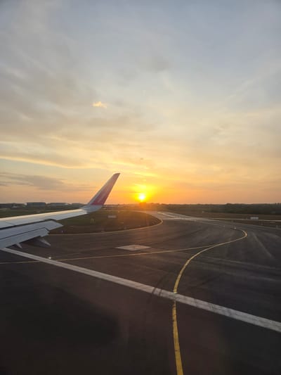 View from the interior of an aircraft during sunrise at an airport. The wing of the aircraft is visible. The aircraft is on the runway, indicated by painted lines. The sky is filled with clouds and the rising sun. The airport is near a forest. The aircraft belongs to the airline Wizz Air. The image does not contain sufficient information to accurately discern the location as North Hertfordshire.