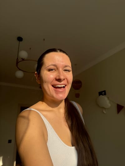 A female individual is depicted in a frontal, chest-up portrait within an interior residential space located in Dubki (Gp Kubinka), Russia. The subject, with dark hair pulled back into a ponytail extending over her right shoulder, wears a white ribbed tank top. She presents a wide, open-mouthed smile, exposing upper and lower dentition, and directly faces the camera. Natural light, originating from the left, strongly illuminates her left side, casting distinct shadows on her right side and the background. The background features a light-colored ceiling from which a dark-framed, circular light fixture with multiple white spherical globes is suspended. The right wall displays a section of red and white triangular bunting and a white cloud-shaped decoration. A dark, rectangular element is partially visible on the far left.