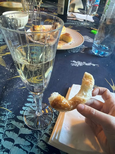 The image shows a table with a dark blue patterned tablecloth. A partially consumed water bottle and a plate with several small pastries are visible in the upper left. A hand holds a curved pastry over a stack of white napkins. A large wine glass lies on its side, partially obscuring the pastries on the plate. The wine glass contains clear liquid, possibly water or white wine. The scene suggests a meal or snack taking place indoors. There is no information to verify if the location is La Lucila, Argentina.