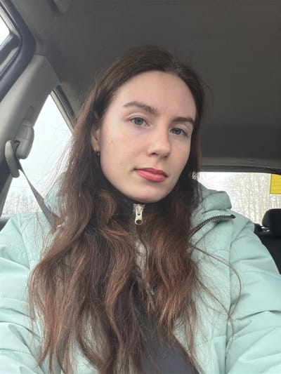 A female subject is depicted seated inside a vehicle, likely in the front passenger or driver's seat. The specific location in Gurlevo, Russia, cannot be determined from the visual evidence. The subject has long, wavy dark brown hair, fair complexion, light-colored eyes, and light red lipstick. She wears a light mint green padded winter jacket with a brass-colored zipper pull visible at the neck, over a dark undershirt or collar. A dark gray seatbelt is fastened across her left shoulder. The vehicle's interior features a gray headliner. Through the side and rear windows, a blurred exterior environment is visible, characterized by bare trees against an overcast, light gray sky. A partial yellow object or sign is visible through the rear passenger window on the right side.