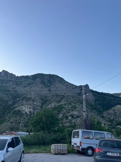 The image shows a mountainous landscape under a clear blue sky. A large rocky mountain dominates the center of the frame, with visible power lines running along its slope. In the foreground, on the right side of the image, are two parked vehicles: a white van and a grey hatchback. Adjacent to the vehicles, there is a stack of paving stones on the side of the road. The image is taken at Bukovo, Bulgaria.