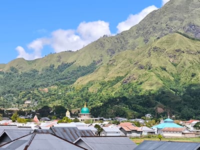 The image captures a wide-angle view of Sembalun Bumbung, Indonesia, dominated by a backdrop of verdant, rolling mountains under a bright blue sky with scattered white clouds. In the foreground, the rooftops of densely packed houses with primarily grey and some red tiling are visible, presenting an aerial perspective.
Mid-ground features include a mosque distinguished by its green dome and a smaller structure with a blue dome. These buildings indicate religious and community spaces within the town. Patches of green fields and trees are interspersed between the houses, suggesting an agrarian landscape.
The scene appears to be shot during the daytime, with good visibility. No people are directly visible within the frame. The overall impression is of a rural Indonesian town nestled in a mountainous region.