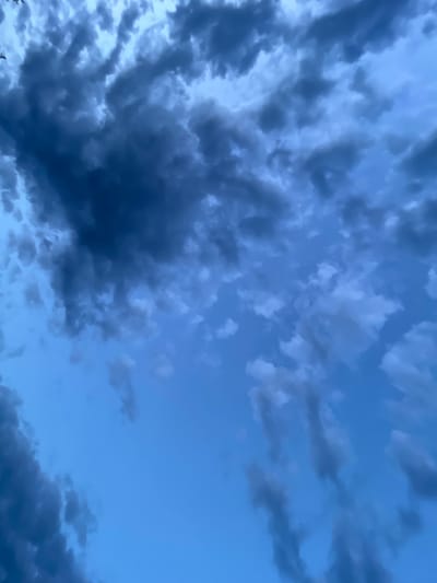 The image shows a close-up of a dark blue sky with scattered clouds. Some of the clouds are dark gray and appear heavy, while others are lighter and more wispy. The sky suggests a possibly stormy or overcast day. There are no people, animals, buildings, or any identifiable landmarks visible in the image. The setting is a sky view, not specific to any particular location within the city of Mayak, Russia.