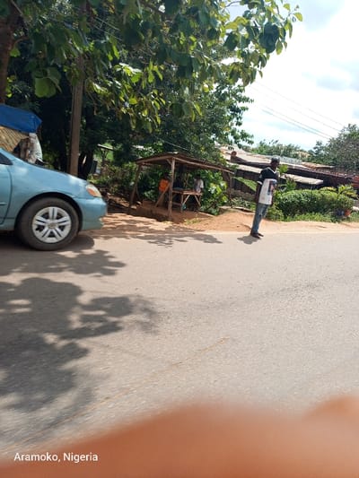 The scene is a paved road in the city of Okinni, Nigeria, located at coordinates 7.828 latitude and 4.517 longitude. A light blue car is parked on the side of the road, facing away from the viewer. There are three people standing to the right of the car, one of whom is standing in the middle of the road. There is a wooden structure, possibly a market stall, in the background. The environment is rural, with trees and vegetation on both sides of the road. The sky is partly cloudy. The image is taken from the perspective of a person sitting or standing in a vehicle. The photo was taken during the day, and the sun is casting shadows on the road.