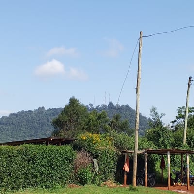 The image depicts a scene in Ihururu, Kenya, on a day with a blue sky and scattered clouds. In the background, a tree-covered hill is visible with several communication towers on its peak. In the foreground is a well-maintained green hedge, partially obscuring a roofed structure. Further to the right, a rudimentary wooden shelter with a corrugated metal roof is supported by wooden posts. Two motorcycles are parked under the shelter. Several people are seated under the shelter. Clothes or fabric items are hanging from the posts of the shelter. Two wooden utility poles with power lines are in the right foreground. The ground is covered in grass with bare earth patches in the shelter area.