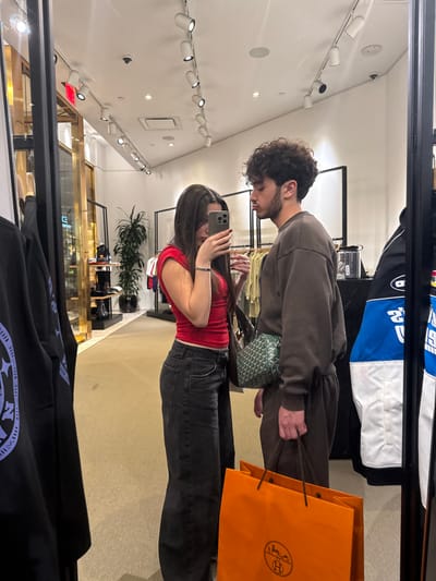 The image depicts a mirror reflection of two individuals inside a modern retail store, located in Winchester, United States of America.

**Scene Description:**
The setting is an indoor commercial space, likely a boutique or clothing store, characterized by a light-colored carpeted floor and a white ceiling equipped with a linear array of white track lighting fixtures. Various clothing items are visible on racks and display units in the background.

**People:**
Two individuals are present in the reflection. On the left, a female with long, dark hair is wearing a red, short-sleeved top and dark grey, wide-leg jeans. She is holding a silver smartphone at chest level, pointed towards the mirror, indicating she is taking the photograph. A dark handbag is partially visible on her right side, and a silver bracelet is on her left wrist. To her right stands a male with dark, curly hair, dressed in a brown long-sleeved sweatshirt and matching brown sweatpants. He has a green patterned fanny pack or crossbody bag worn across his front. His lips are noticeably pursed, and he holds an orange shopping bag with the Hermès logo in his left hand.

**Objects and Environment:**
On the far left, a black garment with a light-colored circular design is partially visible, hanging from a rack. In the background, beyond the individuals, retail displays include a multi-tiered stand with dark footwear or accessories, a tall potted plant, and additional clothing racks. To the far right, another garment, a blue, white, and black jacket or sweatshirt, is visible on a display. The overall lighting is bright and even, originating from the ceiling fixtures.
