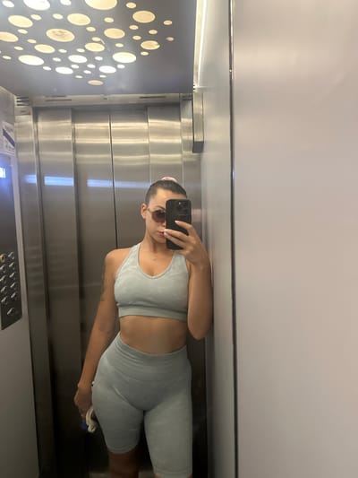 A woman takes a mirror selfie in an elevator. She is wearing a light blue athletic set consisting of a sports bra and high-waisted shorts. She has sunglasses on and her hair is pulled back in a bun with a pink scrunchie. She is holding a phone in her right hand and a blue and white cloth in her left. The elevator has stainless steel walls and a patterned ceiling with circular lights. The floor is not visible. The image was taken in Torremolinos, Spain.