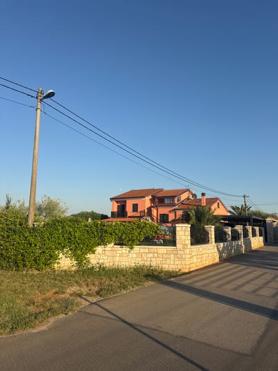 The image depicts a street scene in Crveni Vrh, Croatia, likely during daylight. A residential building, painted in a warm salmon color, dominates the mid-ground. The building features multiple stories, visible balconies with railings, and a terracotta-tiled roof. A palm tree and lush green foliage surround the property, suggesting a warm climate.

In the foreground, a grey asphalt road runs alongside a textured stone wall, which separates the road from the residential property. A street lamp on a utility pole stands to the left, with electrical wires stretching across the clear blue sky. The sky is a vibrant, cloudless blue, indicating fair weather. Shadows cast by the building and the fence suggest the sun is at an angle, possibly late afternoon or early morning.

The scene appears peaceful and residential. There are no people visible in the image. The overall impression is one of a quiet, sunny day in a Mediterranean setting. The image is oriented vertically.