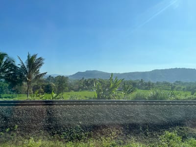 The image depicts a railway track in the foreground, with ballast and vegetation. Beyond the track, a lush green landscape unfolds, featuring rice paddies, palm trees, and other tropical vegetation. In the background, rolling hills are visible under a clear blue sky with a subtle sun flare. The scene is located in the city of Pasiraman Kidul, Indonesia. There are no people or animals visible in the image. The overall impression is one of a serene rural landscape.