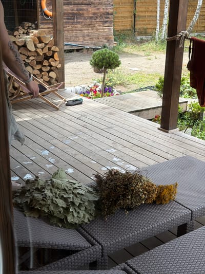 The image depicts an outdoor wooden deck area in Dobrzyń, Poland, adjacent to a rustic wooden building. In the foreground, two sun loungers are covered with dark grey woven material. On the loungers, there are two bundles of dried herbs and foliage, one significantly larger and green, appearing to be a bath whisk (venik), and the other smaller and brown with yellow sprigs. The wooden deck shows some scattered white marks. A person's tattooed arm is visible on the left, with their hand reaching towards a pink and wooden deck chair. Piled firewood is stacked against the wooden building, and a bright orange life preserver is attached to the wall. The background features green grass, a small conical evergreen tree, and various flowering plants. A wooden post with rope tied to it stands on the right side of the deck, with a piece of dark red fabric hanging from it.