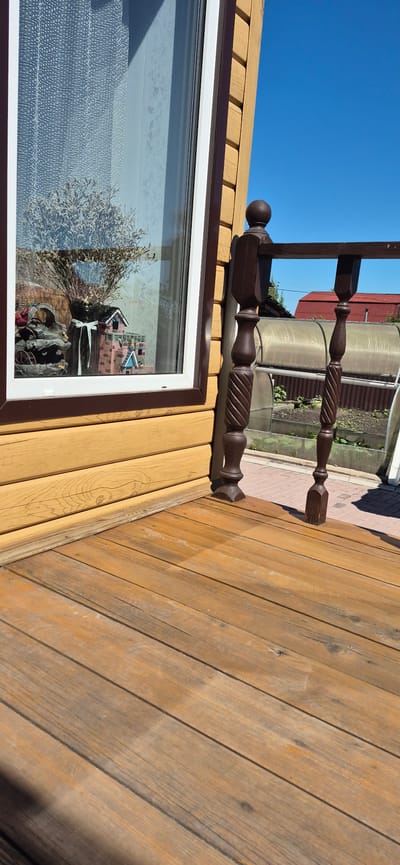 The image depicts a close-up view of a wooden porch with a wooden railing. The porch is constructed from planks of wood with visible grain and some weathering. To the left, a portion of a yellow wooden house with horizontal siding is visible, along with a window. Inside the window, a dried floral arrangement in a vase and a miniature brick house are displayed. To the right of the porch railing, a greenhouse with plants is visible, and beyond that, a red-roofed building. The sky is clear and blue. The scene appears to be outdoors, possibly in a rural or suburban setting, given the presence of the greenhouse and a distant building. There are no people or animals visible in the image. The lighting suggests it is daytime, with bright sunlight casting shadows. The overall impression is one of a sunny, quiet day.