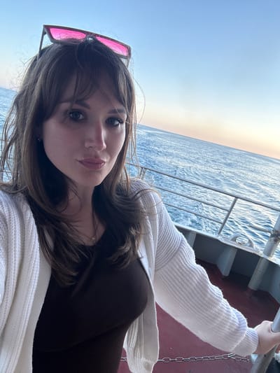 A woman is positioned on what appears to be a ship or boat, holding onto a railing. The railing and ship infrastructure are visible. In the background is the sea and a bright sky. The woman is wearing sunglasses perched on her head, a brown top, and a white knitted cardigan. A gold chain necklace is visible around her neck. The image may have been taken in Гаспринский поселковый совет, Ukraine.