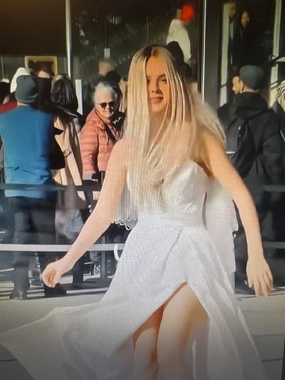 The media file shows a group of people indoors. A woman with blonde hair is in the foreground, wearing a white dress. Her arm is extended, and she appears to be walking or posing. Behind her, several other individuals are visible, some wearing casual clothing and others in darker attire. The setting appears to be a public space, possibly a shopping mall or an event venue, with reflective surfaces and vertical supports suggesting an architectural interior. The lighting is artificial, and there are no clear indications of the time of day or weather conditions. The overall mood is difficult to ascertain due to the limited visibility of facial expressions and the slightly distorted perspective of the image. No visible text is present. The location context provided is Chorzów, Poland.