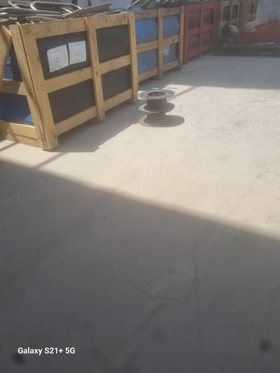 The image shows an outdoor industrial or storage area. In the background, there are several large wooden crates containing blue barrels. Some of these crates are partially obscured by what appear to be metal pipes or rebar stacked on top. To the right of the crates, a person is lying down on a pallet. In the foreground, there is a concrete surface, and a spool of wire is resting on the ground. The area is sunlit, with distinct shadows cast on the concrete, indicating daylight. The bottom left corner of the image contains a watermark that reads "Galaxy S21+ 5G". The location is identified as Pitnak, Uzbekistan.
