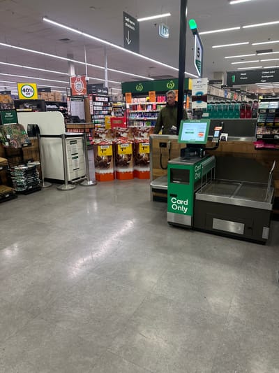 An interior view of a supermarket in Toormina, Australia, shows a self-checkout area with a single customer. The customer, a middle-aged man with short gray hair, is wearing a dark green hooded jacket and is standing at one of the self-checkout machines. He is looking at the touchscreen display, which shows "Welcome" and "Processing." The self-checkout machine is green and black and has "Card Only" printed on its side. It includes a screen, a card reader, and a bagging area. Other self-checkout machines are visible to the left. In the background, shelves are stocked with various products, and overhead signs indicate "Self-serve checkouts" and "Gift Cards." The floor is tiled in a light gray color, and fluorescent lights illuminate the space.