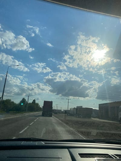 The image depicts a view from inside a vehicle looking forward on a paved road. The road has white lane markings. In the distance, a dark-colored truck is visible, traveling in the same direction. To the right of the road, there are several buildings, some appearing to be industrial or commercial, with parked trucks and trailers. To the left of the road, there are trees and vegetation, as well as utility poles with wires. The sky is bright blue with scattered white cumulus clouds, and the sun is partially obscured by clouds, casting rays of light. The scene suggests an outdoor environment, possibly on the outskirts of a town or city, given the presence of industrial buildings and a highway. The location is identified as Karla Marksa, Russia.