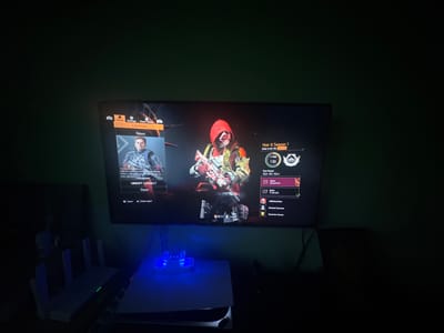 The image shows a television screen displaying the main menu of a video game. The game appears to be a first-person shooter with a post-apocalyptic theme. On the left side of the screen, there is a character model and menu options such as "Continue," "News," and "Ubisoft Connect." On the right side of the screen, there is information about "Year 8 Season 1," including playtime and season level. Below the television, a white gaming console with blue LED lights is visible, along with a black router or modem. The room is dark, with the only significant light source being the television screen and the console's LEDs. There is no visible indication or information within the image that links it to Lesnovo, Bulgaria.