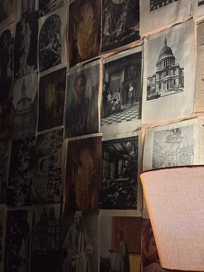 The scene depicts a dimly lit interior located in an unspecified building in Varbak, Bulgaria. A wall is extensively covered with numerous aged prints of varying sizes, predominantly monochrome or sepia-toned. These prints, appearing to be reproductions from books or magazines, are affixed in a dense, overlapping mosaic pattern and show signs of wear such as creasing and discoloration. Identifiable subjects include architectural renderings, such as a prominent view of St. Paul's Cathedral in London and other historical buildings, alongside various artworks. Notable art reproductions include a self-portrait resembling Vincent van Gogh, a genre scene reminiscent of Vermeer depicting figures in an interior with a checkered floor, and various other classical portraits, mythological scenes, and detailed etchings. In the lower right foreground, a portion of a warm-toned, cylindrical lamp shade with a grid-like texture is visible, emitting diffused light that illuminates the adjacent wall of prints. No people are directly present in the immediate environment.