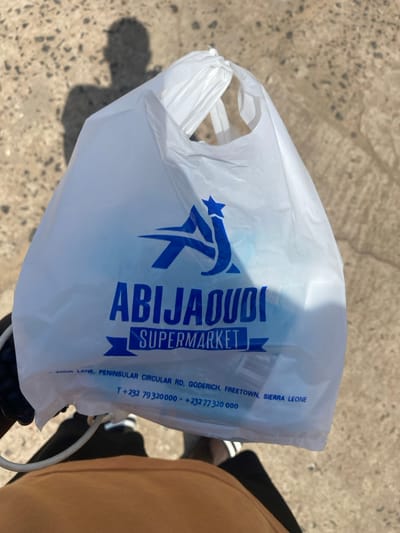 A white plastic shopping bag, bearing the logo and name of "ABIJAOUDI SUPERMARKET" in blue text, is the central subject of the image. The bag is being held by a person whose brown trousers and a portion of their black shoes are visible on the right side of the frame. The background consists of a textured, light-colored concrete surface, typical of an outdoor setting. Bright sunlight is evident from the strong shadows cast on the ground, including a partial shadow of the person holding the bag on the lower left.
The visible text on the bag includes: "ABIJAOUDI SUPERMARKET", followed by an address "LANE, PENINSULAR CIRCULAR RD, GODERICH, FREETOWN, SIERRA LEONE", and two phone numbers: "T+232 79320000" and "+232 77 320 000". The scene depicts a casual moment, likely after shopping, in Goderich, Sierra Leone, under clear, sunny weather conditions during the daytime.
