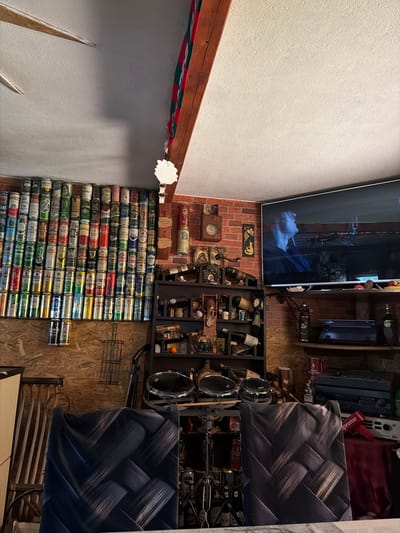 The image depicts an indoor scene that appears to be a room or garage, possibly in Pavolche, Bulgaria. The walls are constructed of wood paneling and brick. A large collection of beer cans is meticulously arranged on shelves and on top of wooden shelving units that line a significant portion of the wall. The cans are of various brands and colors.

Dominating the right side of the frame is a large flat-screen television displaying an image of a person with light-colored hair. Below the TV, there are shelves holding audio equipment, bottles, and decorative items. A dark wooden shelf unit on the left side of the room contains various bottles, including wine bottles, and other objects, some of which appear to be decorative figurines and religious icons.

In the foreground, two chairs with patterned dark upholstery are visible. Behind the chairs, a drum set with two cymbals and two drums is set up. Above the beer cans, a wooden beam with a red, green, and white striped fabric draped over it is present, attached to the ceiling. The ceiling itself is textured and appears to be white or off-white, with a visible ceiling fan on the left side. The overall impression is of a personal space decorated with a passion for beer and potentially music, with a mix of organized displays and accumulated items.
