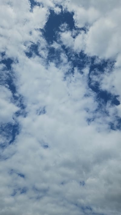 A full-frame vertical shot captures a sky filled with scattered white clouds against a deep blue background. The clouds are varied in shape and density, with some appearing wispy and translucent, while others are more solid and opaque. Patches of clear blue sky peek through the clouds in several areas. The lighting suggests it is daytime, possibly mid-morning or afternoon, given the bright blue of the sky and the illumination of the clouds. There are no identifiable landmarks, people, or objects within the frame, indicating the image focuses solely on the atmospheric conditions. The scene depicts a natural sky, likely captured from an open area in the city of Kroņi, Latvia, with no specific points of reference to indicate a precise location within the city.