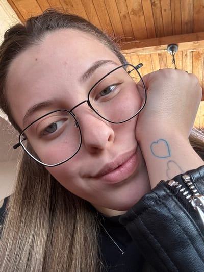 A close-up image depicts a person with light skin tone, brown hair, and greenish-grey eyes, wearing black-rimmed glasses. The person is looking slightly upward and to the right of the frame with a subtle smile, resting their left hand against their cheek. Two outlined heart tattoos, one blue and one faded grey, are visible on the inner left wrist. The person is wearing a black garment and a black faux leather jacket with a silver zipper on the right arm. A thin silver necklace is also visible. The background consists of a wooden slatted ceiling, from which a dark-colored light fixture or hook is partially visible in the upper right. The setting appears to be indoors. The precise location within Dimitrovgrad Municipality, Serbia, cannot be determined from the provided image.