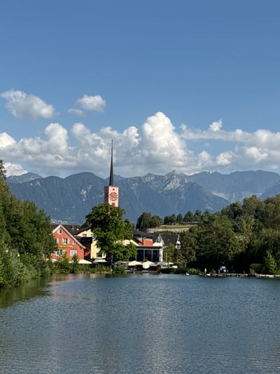 This is a photograph of a scenic lake in Buchs (SG), Switzerland. In the foreground, a calm body of blue water reflects the sky and surrounding scenery. On the far bank of the lake, a cluster of buildings is visible. One prominent building is red with white trim and windows. Next to it, a yellow building can be seen. A tall, slender clock tower with a pink face and a dark spire rises above the buildings, its hands indicating the time. Behind the buildings, a dense line of green trees creates a natural barrier. Further in the background, a range of imposing, blue-tinted mountains stretches across the horizon, their peaks partially obscured by the haze of distance. The sky above is a bright blue, dotted with fluffy white clouds. Along the edge of the lake, several white umbrellas are set up, suggesting an outdoor seating area for a cafe or restaurant. A few individuals are present near the water's edge, with two white swans also visible.