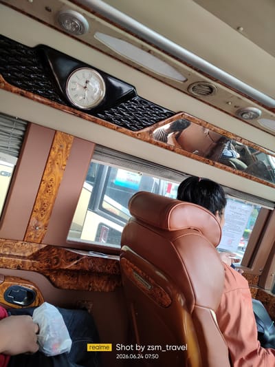A person is seated on a brown leather bus seat, facing away from the camera and looking out a window. The bus interior features wood-grain trim and a clock on the ceiling. Outside the window, another bus is visible. A plastic bag with items inside is held in the person's lap. A timestamp and watermark are visible in the lower right corner of the image, indicating it was taken on June 24, 2026, at 07:50, and shot by zsm_travel. The location is identified as Gò Dầu, Vietnam.