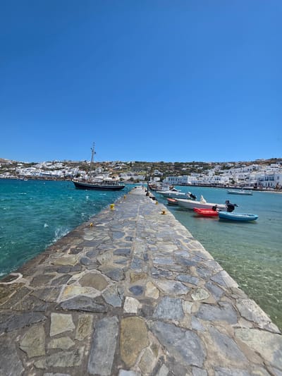The image depicts a scenic view of the harbor in Mykonos Town, Greece, under a clear blue sky. A stone pier, constructed from irregular grey flagstones, extends into the turquoise waters of the Aegean Sea. Along the pier, several yellow bollards are visible, used for mooring boats.
In the foreground, on the right side of the pier, several small boats are moored. These include a bright red kayak, a blue dinghy, and two white motorboats. The water around the pier is exceptionally clear, allowing the seabed to be visible in some areas. On the left side of the pier, a larger, traditional wooden boat with a tall mast is anchored, its hull painted dark blue and white.
The background showcases the iconic white-washed buildings of Mykonos Town, cascading down a hillside towards the harbor. The architecture is characterized by cubic structures with blue accents, typical of the Cycladic style. The town appears to be bustling, with some figures visible along the waterfront promenade. The sea is calm, with gentle waves lapping against the pier. The overall scene conveys a serene and picturesque Mediterranean atmosphere.